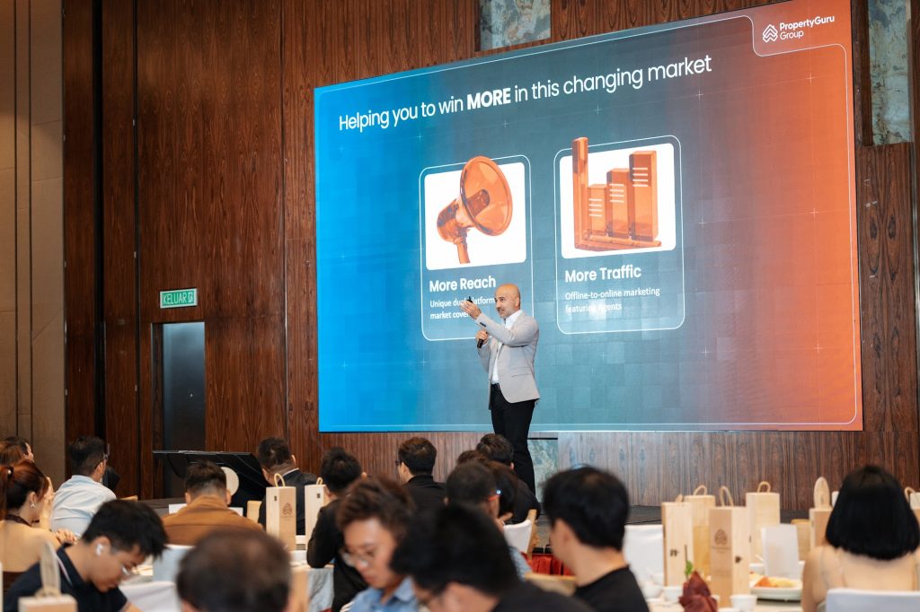 Speaker presenting to seated guests at a corporate seminar with slides on a large LED screen